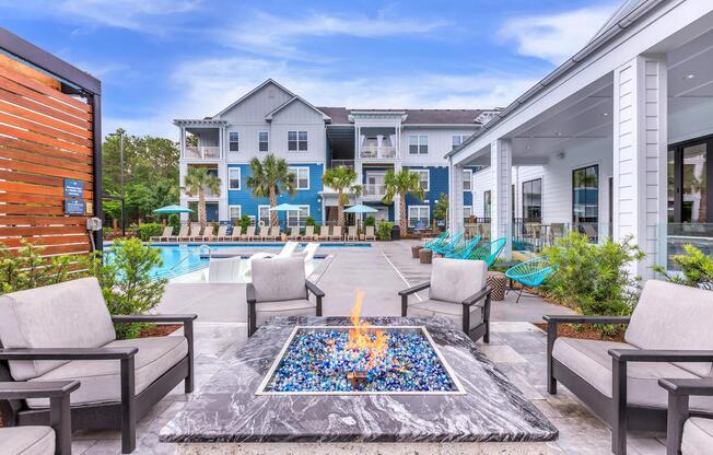 A modern outdoor lounge area featuring a fire pit with blue glass stones, surrounded by four gray chairs. In the background, there are palm trees, a swimming pool, and a stylish apartment building with multiple stories, all set against a bright blue sky.