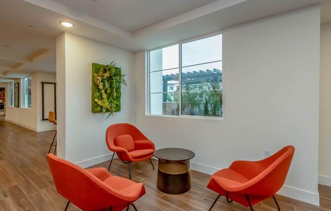 A modern interior space featuring two bright orange chairs and a small round table, with a large window allowing natural light. A green wall art piece adds a touch of nature to the neutral-colored walls, while additional seating is visible in the background. The flooring is made of wooden planks.
