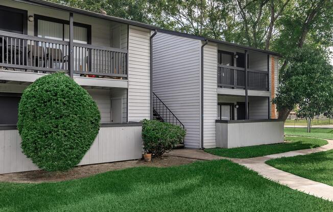 Two adjacent apartment buildings with a light gray exterior, featuring balconies and shrubs. A curved walkway leads through a well-manicured green lawn, with trees in the background.