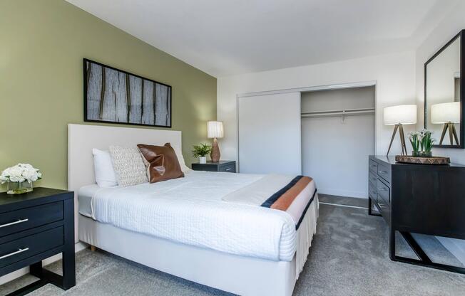 A cozy bedroom featuring a neatly made bed with white linens and a brown throw pillow. There is a nightstand with a lamp and a decorative plant. A minimalistic artwork hangs on the green wall above the bed. A closet is visible in the background, along with a dark dresser displaying a small bouquet of flowers.