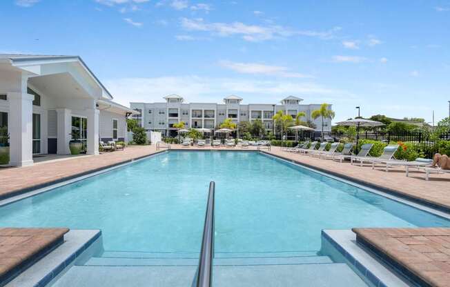 A large swimming pool in front of a white building with lounge chairs around it.