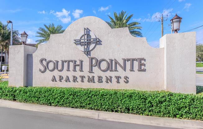 Sign for South Pointe Apartments, featuring the name prominently displayed. The sign is set against a backdrop of blue sky and green landscaping, including palm trees and manicured bushes, indicating a welcoming residential community.