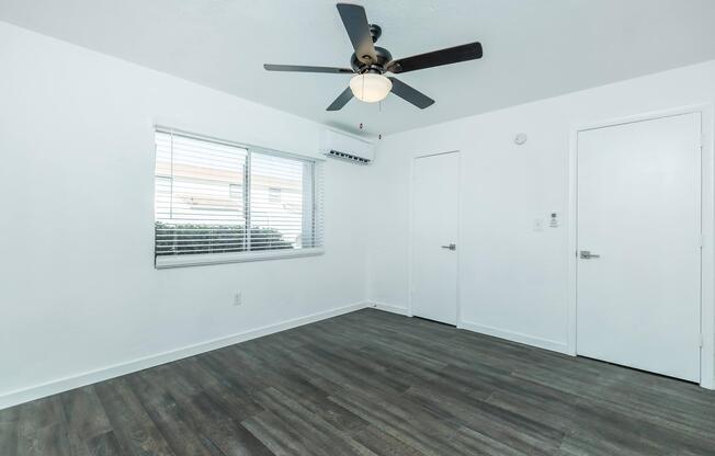 A bright, empty room with white walls and a light wood floor. It features a ceiling fan with light, a window with blinds providing natural light, and two white doors leading to adjoining spaces. An air conditioning unit is mounted above the window.