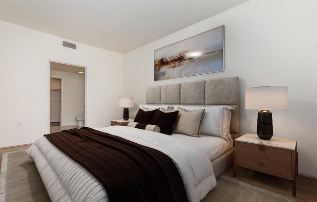 A cozy bedroom featuring a large bed with plush pillows and a dark throw blanket. The room includes two bedside tables with modern lamps, neutral-toned walls, and a piece of abstract art above the headboard. A doorway leads to a bathroom, visible in the background. Soft lighting creates a warm ambiance.