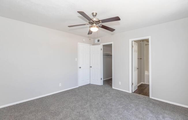 A spacious, neutral-toned bedroom featuring a ceiling fan, light gray walls, and plush carpet. Two doors lead to a closet and a bathroom, respectively. The room has a bright, airy feel, with natural light enhancing the space. Ideal for personalization or as a cozy retreat.