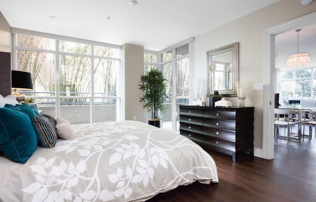 One-Bedroom Apartments in Irvine, CA - Astoria at Central Park West - Bedroom with Dark Wood-Tone Vinyl Flooring, Bed, Wooden Dresser, Framed Mirror, Lamps, House Plant, and Large Floor-to-Ceiling Windows.