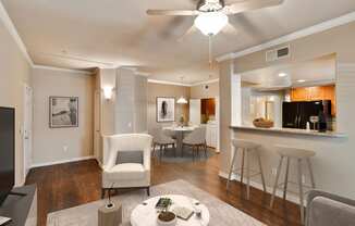 Photo of a model apartment living room and kitchen bar stool area with dining area in the background. The coffee table and white chair can be seen in the living room with bar stools facing the kitchen. A round dining table with four chairs is in the dining room.
