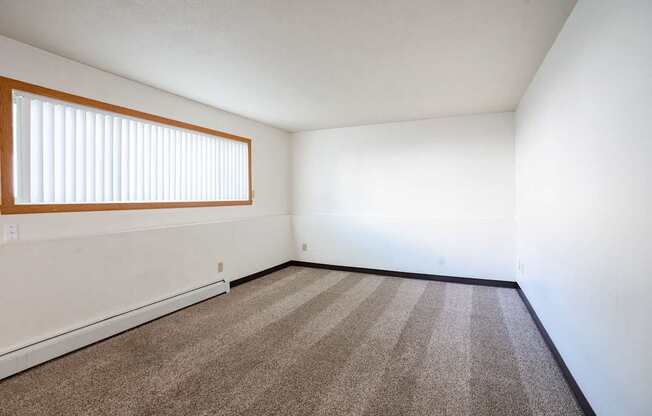 A room with a carpeted floor and a window with blinds. Fargo, ND Islander Apartments