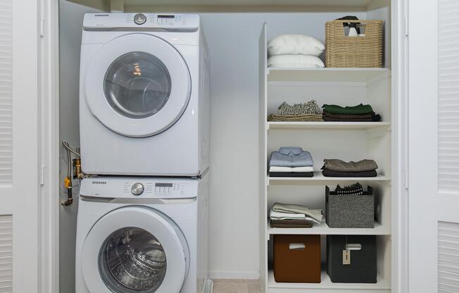 A stacked washer and dryer in a laundry closet, next to shelves filled with neatly folded towels and various storage baskets. The space is well-organized and designed for efficient use, featuring a clean and modern aesthetic.