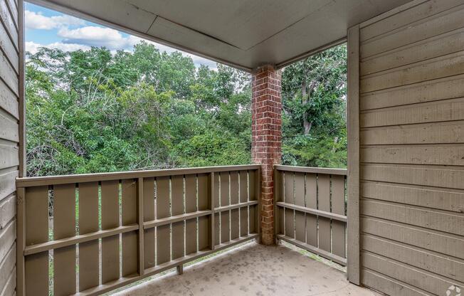 View from a balcony overlooking a green landscape. The balcony features rustic wooden railings and a ceiling, surrounded by trees and foliage in the background. The scene conveys a peaceful outdoor setting, ideal for relaxation.