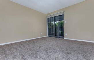 Empty living room with light beige walls and a large window covered by vertical blinds. The floor is carpeted in a soft brown hue, and there are no furniture items in the room, creating a spacious and open feel. Natural light filters through the window, illuminating the area.