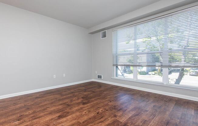 A spacious, empty room with light wood laminate flooring and a large window covered with blinds. The walls are painted in a neutral gray color, and there's natural light coming through the window, illuminating the space.