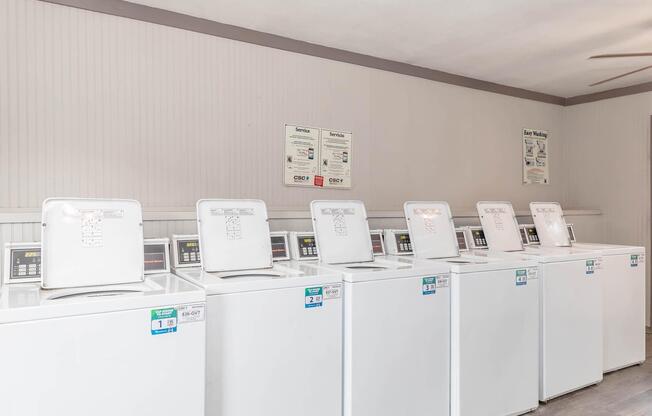 A row of six white washing machines in a laundry room, each with digital control panels. The room has light-colored walls and a ceiling fan, with informational signs posted on the wall above the machines. The space appears clean and well-maintained.