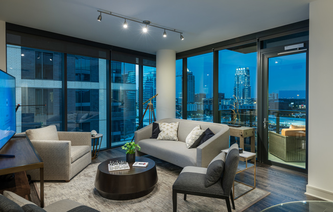 Spacious living room with two walls of full-width floor-to-ceiling windows allowing for a panoramic city view. The room has a couch, coffee table, three armchairs, media console, dark wood-style floors, and a door to a balcony.