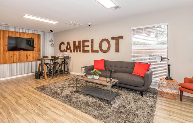 Cozy living room area featuring a gray sofa with red cushions, a wooden coffee table, and a decorative rug. The wall displays the word "CAMELOT" in large wooden letters. A flat-screen TV is mounted on the wall, and there are stylish accents like plants and a modern chair, creating a welcoming atmosphere.