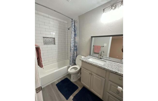 A modern bathroom featuring a white tiled shower with a glass accent, a light gray wall, a countertop with a sink, and a large mirror. The floor is covered with light gray vinyl, and there are two dark blue bath mats. A toilet is adjacent to the sink, with a towel hanging nearby.