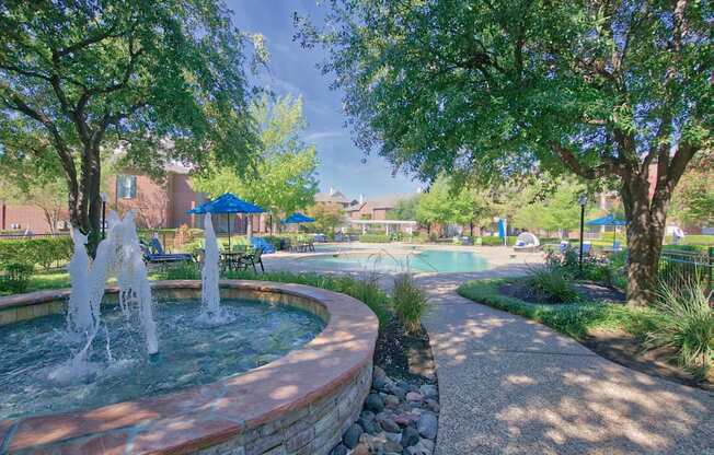 A fountain in the middle of a park with trees and a pool in the background.