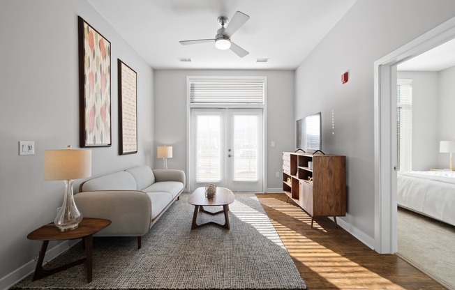 A living room with double doors to a patio providing natural light, hardwood-style flooring, a ceiling fan, and access to a bedroom.