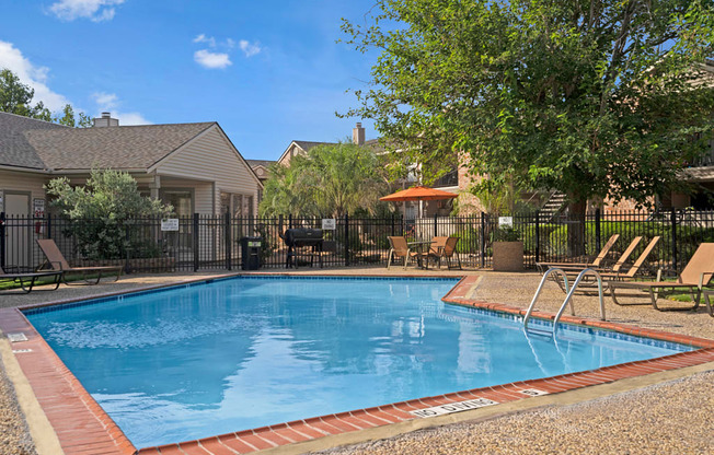 A bright outdoor swimming pool here at Northridge Court featuring clear blue water, a surrounding textured pool deck with lounge chairs, a safety handrail, and shaded seating areas with umbrellas, framed by mature trees, landscaped greenery, and nearby apartment buildings under a sunny blue sky.