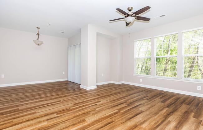 A spacious room featuring a light-colored wall, large windows with blinds, and a ceiling fan. The floor is made of polished wood, giving the space a warm appearance. There is a light fixture hanging from the ceiling, and a sliding door is partially visible on the left side, leading to a potential outdoor area.