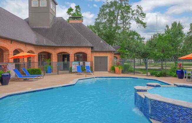A tranquil outdoor swimming pool surrounded by lounge chairs and vibrant orange umbrellas. The pool features a stone waterfall and is located near a brick building with large windows. Lush green trees and a well-maintained lawn are visible in the background under a partly cloudy sky.