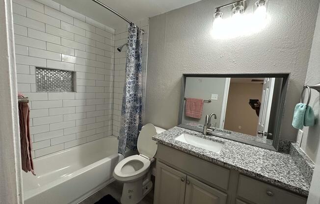 A modern bathroom featuring a white tub and shower with tile accents. There's a light gray vanity with a granite countertop and a mirror above it. Towels hang nearby, and a window provides natural light. The walls are painted a neutral color, creating a clean and inviting atmosphere.