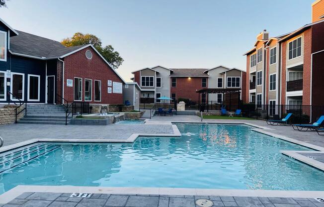 A clear swimming pool surrounded by lounge chairs, with a few trees in the background. On one side, there are modern brick and siding apartment buildings. The setting is tranquil, indicating an outdoor recreational area, likely in a residential complex. The sky is clear and the lighting suggests early evening.