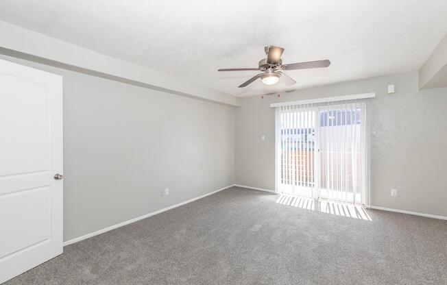 Spacious interior of a room featuring light gray walls, plush carpet, a ceiling fan, and sliding glass doors leading to an outdoor area. Natural light enters the room, enhancing its inviting atmosphere. The door is closed, and there are no visible furnishings.