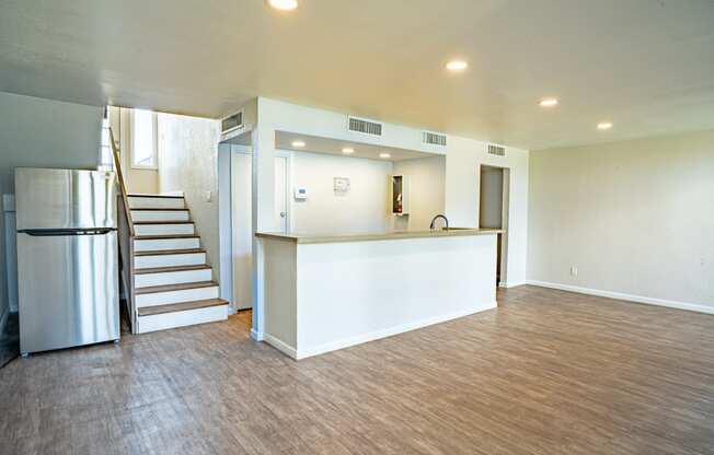 an empty living room with a kitchen and a stainless steel refrigerator
