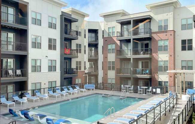 A large swimming pool surrounded by lounge chairs in front of apartment buildings.