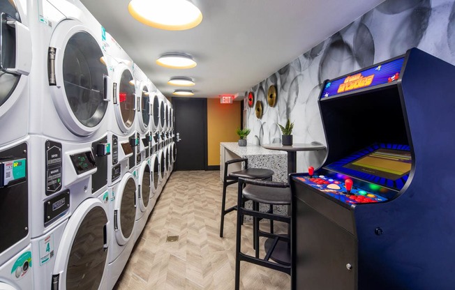 A row of washing machines are lined up in a room with a pinball machine and a bar stool.