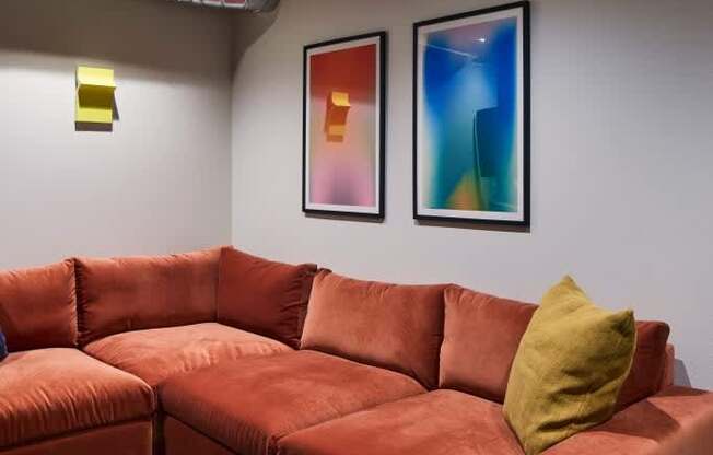 A red couch with a yellow pillow is in the center of a room with two framed pictures on the wall.