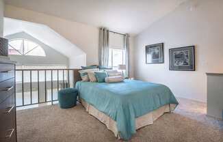 A bedroom here at Lakewood on the Trail with soft carpet, a teal bedspread and matching accent pillows, a small teal ottoman, and bright windows near a loft-style railing, creating a fresh and inviting sleeping space.