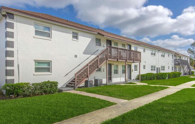 A multi-unit residential building with a white exterior, featuring two levels and wooden staircases. The building is surrounded by well-maintained green lawns and shrubs, with a paved walkway leading up to the entrance. The sky is partly cloudy, creating a bright atmosphere.