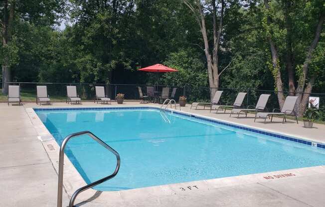 Rectangle pool with brown lawn chaairs, table with 4 chairs and red umbrella in back corner