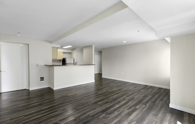 An Unfurnished living room with dark plank flooring, recessed lighting, and tons of space with a view of the kitchen in the background at Arabella Apartment Homes, WA 98155