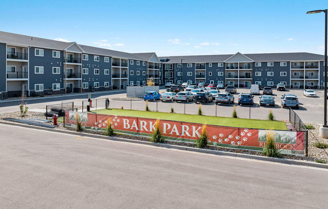 A parking lot in front of apartment buildings with a sign that says Bark Park. at Red Rock Apartments, South Dakota