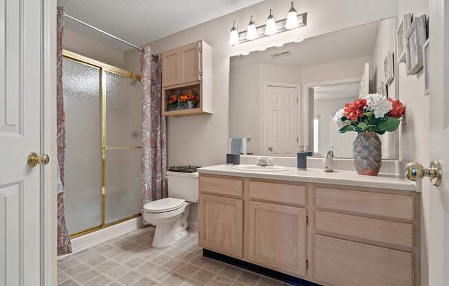 A bathroom with a sliding glass door shower, a linen closet, light wooden cabinets over the toilet, and a vanity with a large mirror and storage under the sink.
