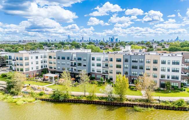 A large building complex sits next to a body of water with a city skyline in the background.