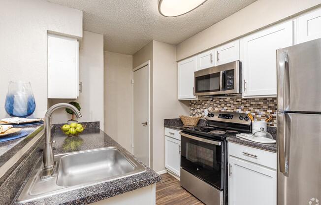 A modern kitchen featuring white cabinetry, a stainless steel refrigerator, a microwave, and a black stove. The counter is dark granite, and there's a stainless steel sink. Decorative items, including a blue vase and a bowl of fruit, add a touch of color. The walls are light-colored, creating a bright and inviting atmosphere.