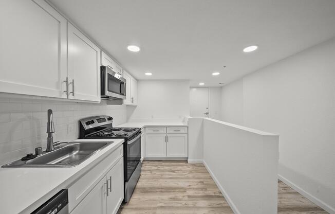 Modern kitchen with white cabinetry, stainless steel appliances, and a single basin sink. The layout features a breakfast bar and a light, airy atmosphere with neutral-colored walls and wood-like flooring. Bright recessed lighting enhances the spacious feel of the area.