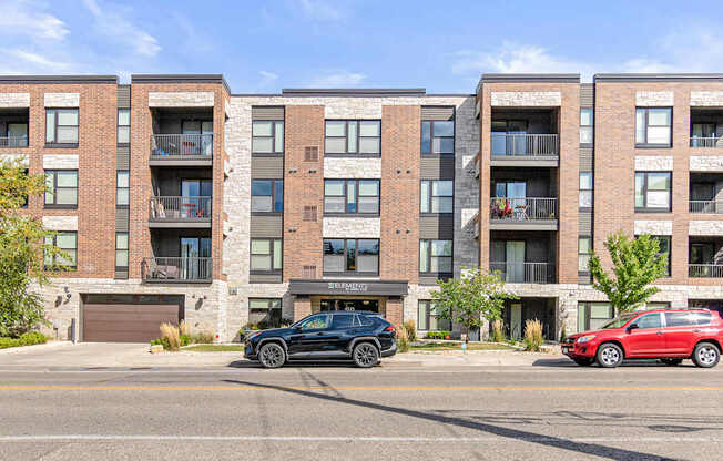 A black car is parked in front of a red car in front of a brick building.