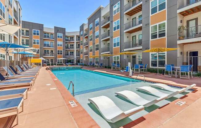 a swimming pool with chaise lounge chairs and umbrellas in front of an apartment building