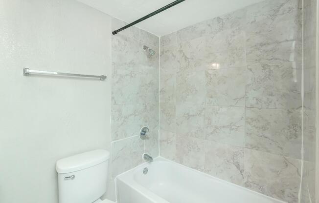 A clean bathroom featuring a white toilet, a bathtub with a showerhead, and light-colored marble walls. A towel bar is mounted on the wall, and the overall space is bright and uncluttered.