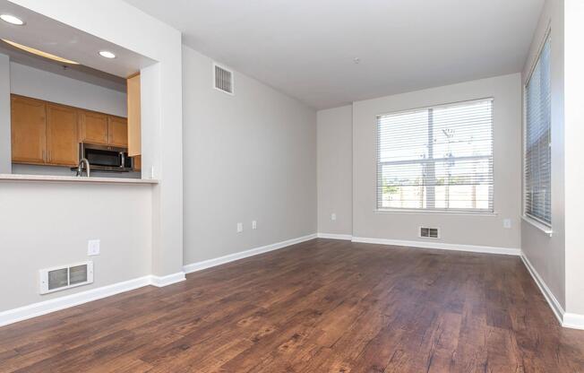 A spacious and empty living area with hardwood flooring, featuring a large window with blinds, light-colored walls, and a partial view of a kitchen with wooden cabinetry and appliances. The room has a contemporary feel, creating an inviting atmosphere for potential furnishings and decor.