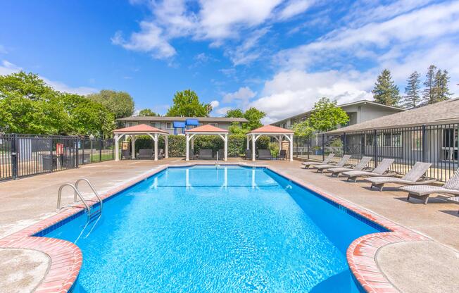 A clear blue swimming pool surrounded by lounge chairs and shaded seating areas. The pool is enclosed by a black fence, with green trees and a cloudy sky in the background, creating a relaxing outdoor atmosphere.