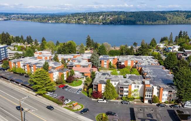 A scenic aerial view here at Watercrest showcasing well-maintained apartment buildings, landscaped grounds, and ample parking, set near a beautiful waterfront with expansive water and tree-lined views, creating a peaceful and desirable location.