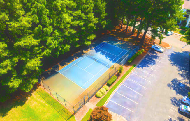 A tennis court surrounded by trees and a parking lot.