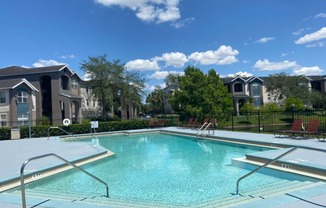 Community pool with sundeck, lounge chairs, surrounded by black metal fence with trees, lake, and building exteriors in the background
