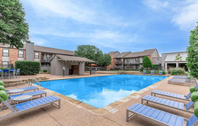 A spacious outdoor swimming pool surrounded by lounge chairs and greenery, with residential buildings in the background. There are shaded seating areas and a clear blue sky overhead, creating a relaxing leisure atmosphere.
